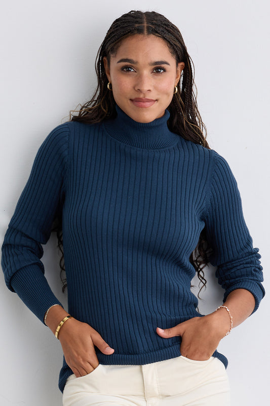 A woman with long braided hair wears a blue ribbed turtleneck sweater and cream pants, standing against a light-colored wall with her hands in her pockets.